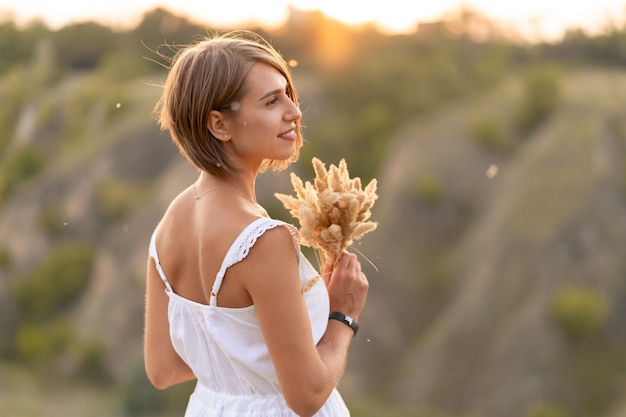 Linda garota concurso em um vestido branco caminha ao pôr do sol em um campo com um buquê de espiguetas.