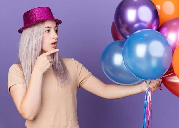 Linda garota confusa usando chapéu de festa segurando e apontando para balões