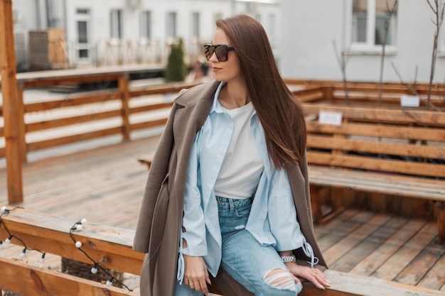 Linda garota da moda com óculos de sol vintage em uma roupa casual da moda com uma elegante camisa de casaco e jeans senta-se e descansa em uma cerca de madeira na rua