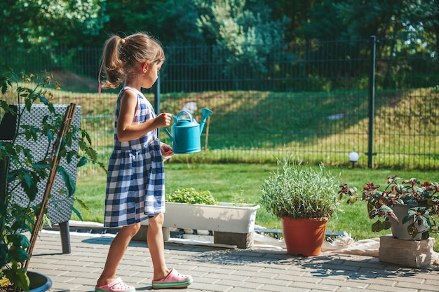 Linda garota de 3 a 4 anos de idade em um vestido xadrez azul regando as plantas de um regador no jardim. Crianças de atividades ao ar livre.