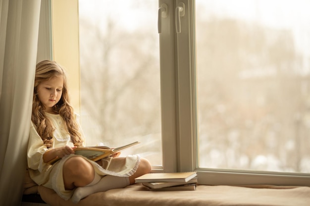 Linda garota de cabelo comprido lê um livro sentado no parapeito da janela