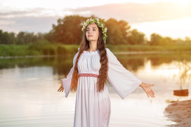 Foto linda garota de cabelo preto com vestido vintage branco e coroa de flores em pé na água do lago. reflexo do sol.