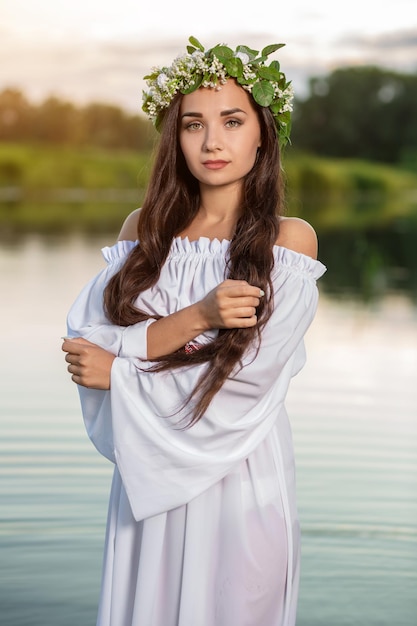 Linda garota de cabelo preto com vestido vintage branco e coroa de flores em pé na água do lago. Reflexo do sol.
