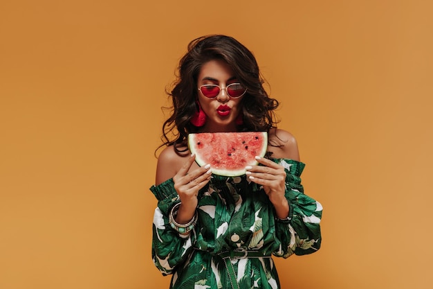 Linda garota de cabelos encaracolados com óculos de sol modernos em vestido verde impresso de verão posando com melancia em pano de fundo laranja