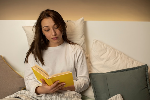 Foto linda garota de cabelos escuros de pijama lendo livro com capa amarela, sentado na cama