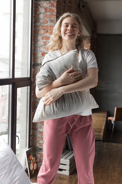 Linda garota de calça de pijama rosa e camiseta branca em pé na cama com travesseiro e sorrindo