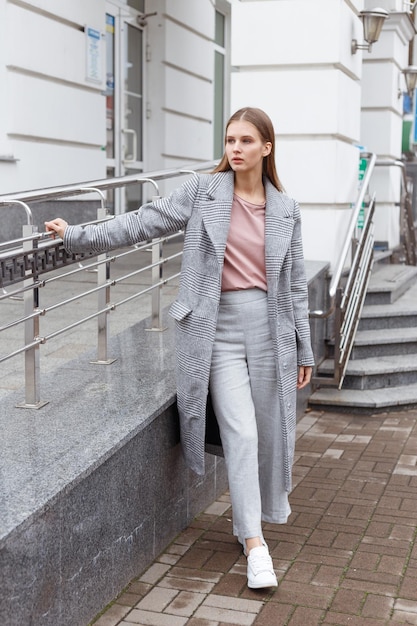 Linda garota de casaco cinza na moda e calças sobre fundo claro andando na rua