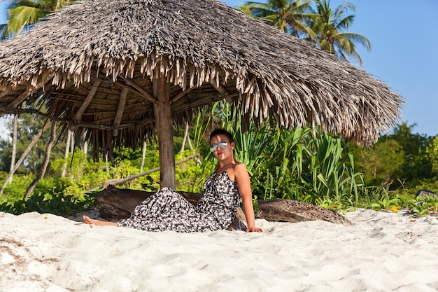 Linda garota de macacão e óculos escuros, senta-se sob um guarda-sol com bengala na praia com palmeiras altas e areia branca. Costa Africana do Oceano Índico de Mombaça