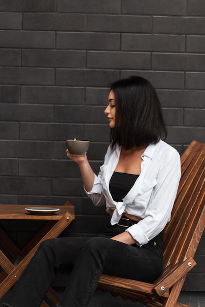 Linda garota de negócios em uma camisa branca da moda com jeans preto senta-se em uma cadeira de madeira e bebe café aromático perto de uma parede de tijolos pretos na rua