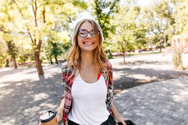 Linda garota de óculos e fones de ouvido passando tempo no parque foto ao ar livre de encantadora senhora caucasiana em roupa de primavera em pé no fundo da natureza com sorriso