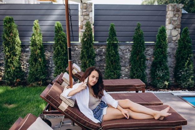 Linda garota de pé em um fundo da piscina e falando ao telefone ela está feliz Ela está vestida com uma camiseta branca elegante com uma estampa de palmeiras mochila azul óculos azuis