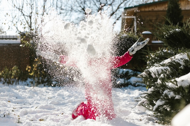 Linda garota de sete anos com roupas de inverno, brincando com neve no quintal de uma casa em um dia ensolarado de inverno.