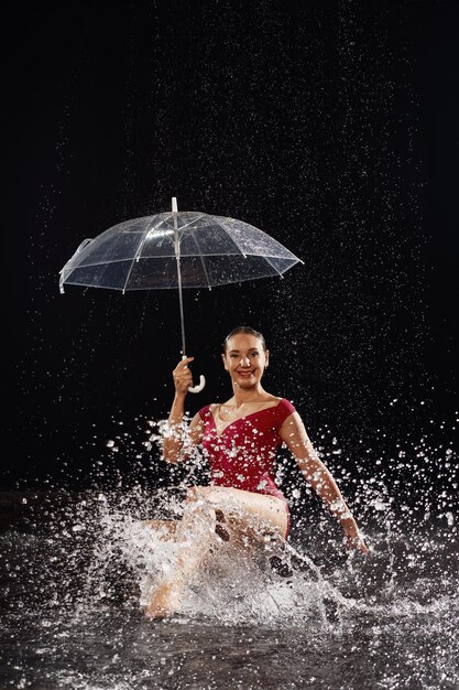 Linda garota de sportswear vermelho com guarda-chuva no estúdio aqua Gotas de água espalhadas sobre seu corpo de fitness A figura perfeita no fundo de salpicos de água Fundo clássico de cor escura