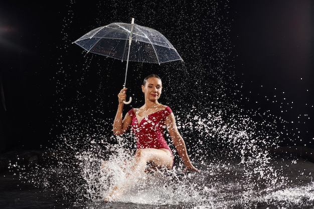 Linda garota de sportswear vermelho com guarda-chuva no estúdio aqua Gotas de água espalhadas sobre seu corpo de fitness A figura perfeita no fundo de salpicos de água Fundo clássico de cor escura