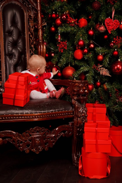 Linda garota de um ano de idade no vestido de Papai Noel no trono no fundo da árvore de Natal decorada brinquedos. Criança em decorações com caixas de presente. Conceito de celebração do ano novo em casa aconchegante. Copie o espaço