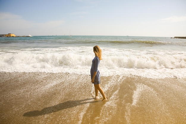 Linda garota de vestido azul está andando na praia Incrível foto de verão Mulher perto do mar Conceito de viagem de férias Pernas finas Água quente do oceano