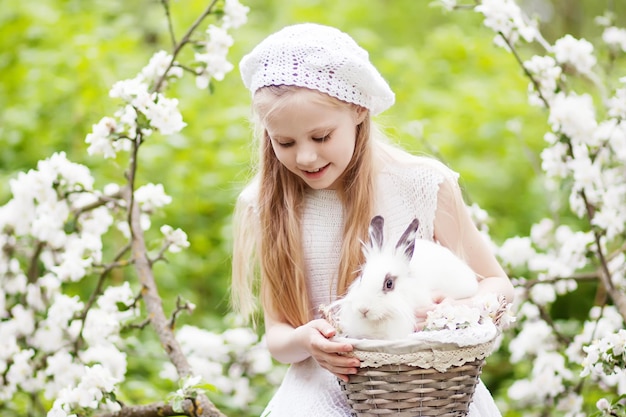 Linda garota de vestido branco brincando com coelho branco no jardim de flores da primavera Atividade divertida de primavera para crianças tempo de páscoa