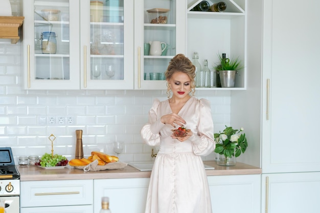 Linda garota de vestido lindo come morangos maduros na cozinha Luxuoso