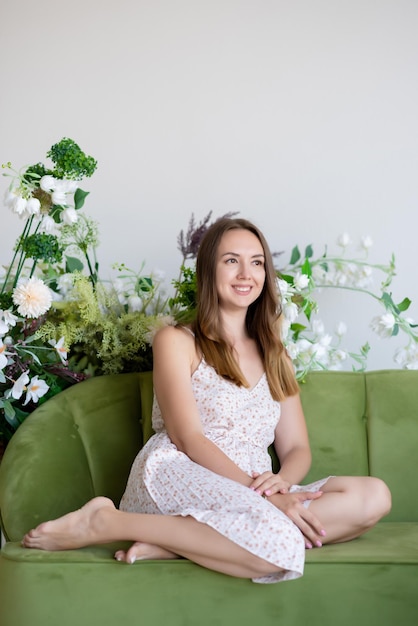 Linda garota de vestido senta-se em um sofá verde em uma sala cercada por flores sorrindo olhando para longe