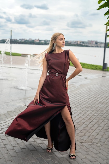 Linda garota de vestido vermelho escuro longo e elegante perto da lagoa no parque sity