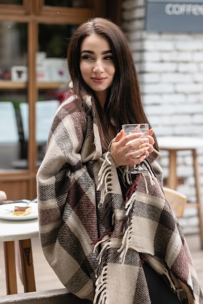 Linda garota descansando e bebendo café sentado em um café no terraço, envolto em um cobertor xadrez de lã. Manhã de outono. Mulher jovem e bonita com uma xícara de café ao ar livre em dia de outono