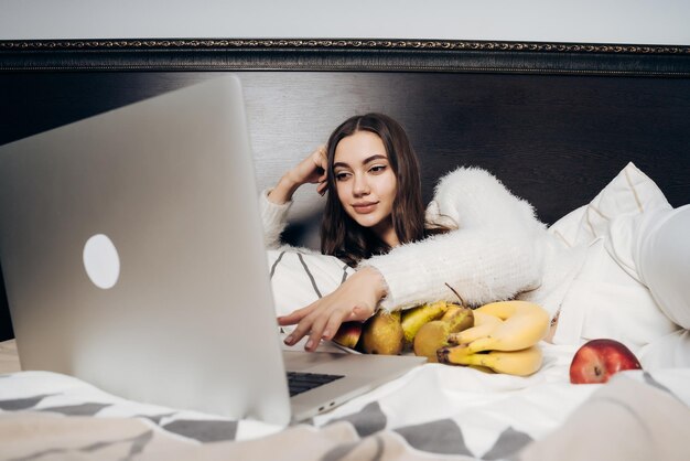 Linda garota descansando na cama depois de um árduo dia de trabalho assistindo a um filme interessante no laptop e comendo frutas