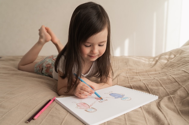 Linda garota desenha um desenho a lápis. uma criança aprende a desenhar pessoas. a criança deita na cama e desenha com um lápis azul