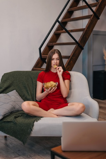 Foto linda garota doméstica comendo batatinhas, assistindo tv, sentado no sofá.