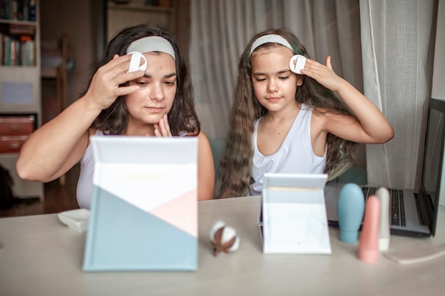 Linda garota e a mãe dela assistindo aula de beleza com tablets online e fazendo procedimentos de spa sozinhas, mãe e filha se divertindo com máscara lisa no rosto, salão de beleza em casa