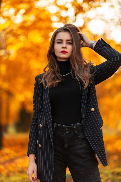 Linda garota elegante com lábios vermelhos em roupas da moda com uma jaqueta, blusa e calça jeans preta caminhando contra um fundo de folhagem de outono laranja brilhante ao pôr do sol