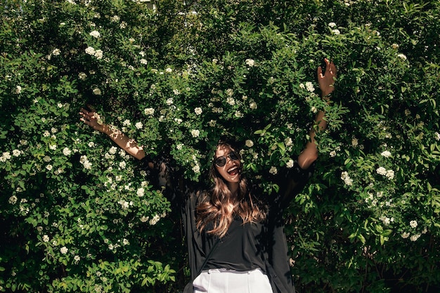 Linda garota elegante Hipster com cabelo lindo relaxando em arbustos florais em dia ensolarado Mulher Boho feliz com óculos de sol em roupa da moda, aproveitando o dia em flores no jardim