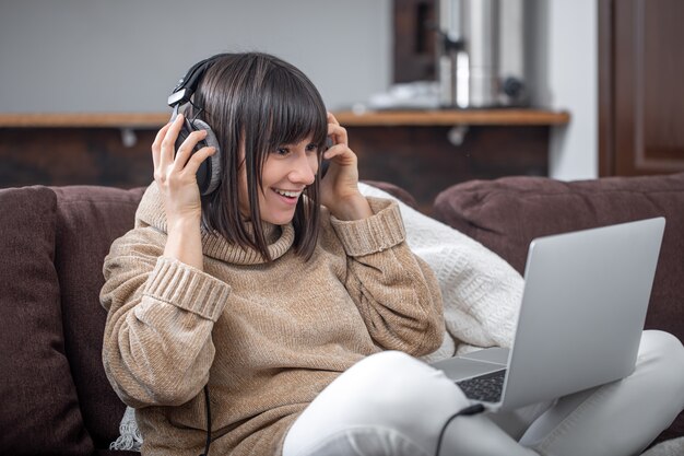 Linda garota em fones de ouvido, ouvindo música em casa no sofá com um laptop.