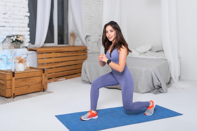 Linda garota em um agasalho roxo está fazendo fitness em casa Garota fitness fazendo exercícios em um tapete de ioga Uma garota em forma levando um estilo de vida ativo Sorrindo olhando para a câmera