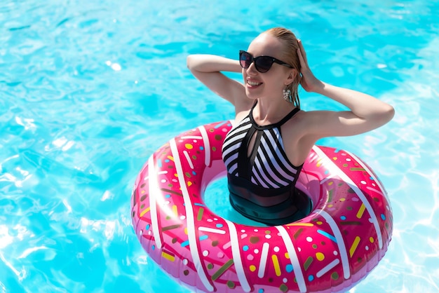 Linda garota em um maiô preto e branco e óculos de sol nada na piscina com um círculo inflável na forma de um donut gosta de relaxar na piscina em um dia quente e ensolarado