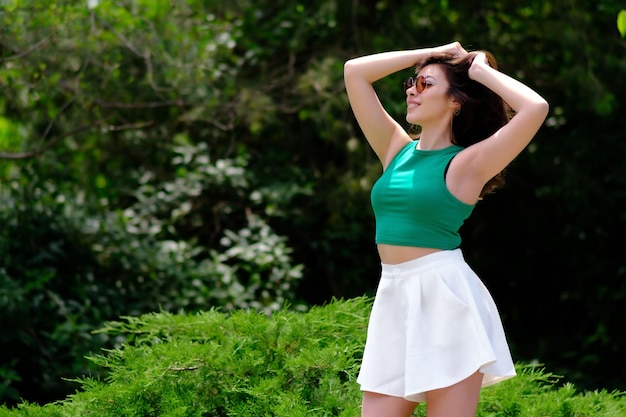 Linda garota em um top verde e shorts brancos em um parque da cidade em um dia ensolarado de verão Posando com as mãos segurando o cabelo