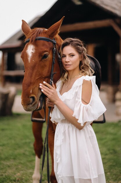 Foto linda garota em um vestido branco ao lado de um cavalo em um antigo rancho