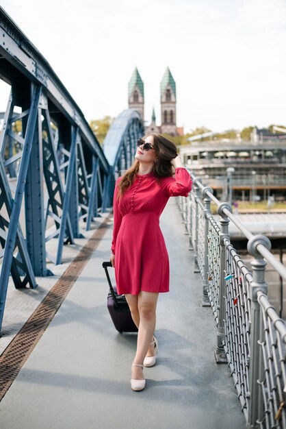 Linda garota em um vestido vermelho com uma mala indo ao longo da ponte olhando para cima e admirando uma nova cidade