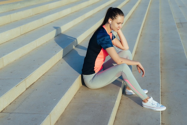 Linda garota esportiva descansando na escada depois de uma corrida, ouvindo música em fones de ouvido