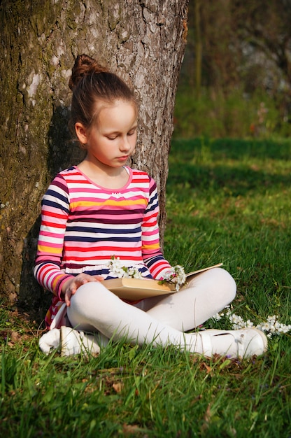 linda garota está lendo o livro no parque, sentado na grama perto da árvore