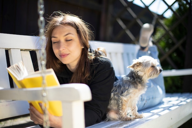 Linda garota está lendo um livro enquanto estava deitado em um banco com um cachorro em um dia ensolarado. descanse e relaxe no conceito de jardim. recreação ao ar livre. férias na aldeia. dia quente de verão ensolarado.