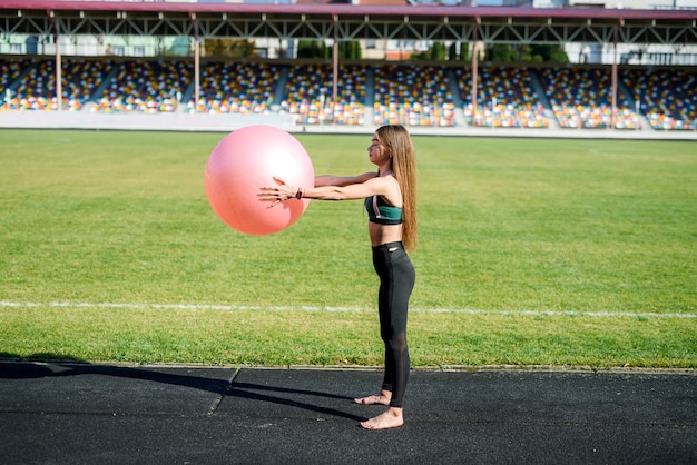 Linda garota fazendo exercícios no fitball no estádio em um dia ensolarado. Estilo de vida saudável na cidade