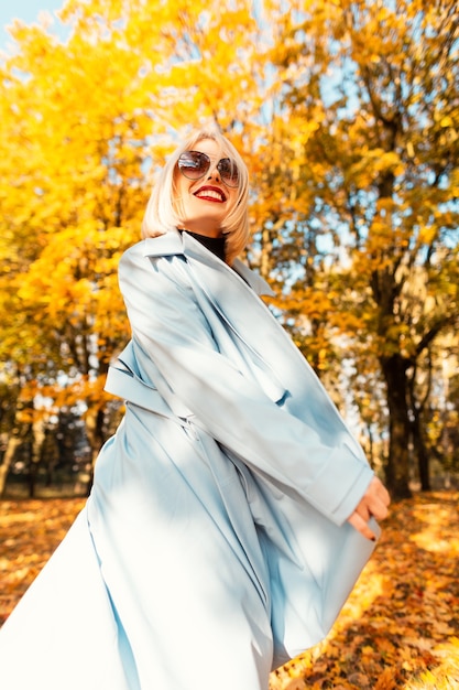 Linda garota feliz com um sorriso em um casaco azul da moda caminha em um parque de outono com folhagem amarela brilhante em férias ensolaradas
