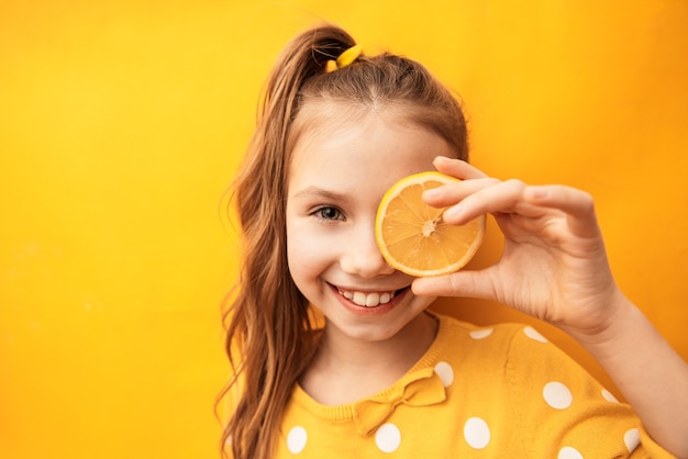 Linda garota feliz com uma pele limpa e perfeita, escondendo o olho atrás de uma fatia de limão em fundo amarelo