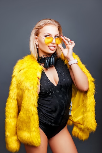 Linda garota feliz e elegante com fones de ouvido em elegantes óculos de sol amarelos e um elegante casaco de pele amarelo na cor cinza