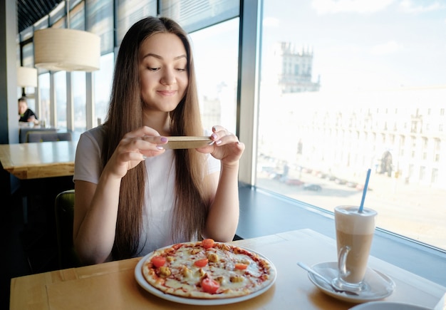 Linda garota feliz está fazendo foto de comida no café, café com leite na pizza de mesa, comunicação nas redes sociais