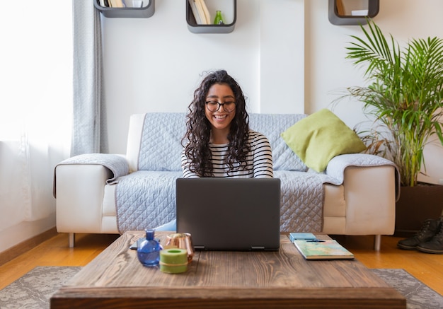 Foto linda garota feliz está sentada e usa seu computador cinza atrás dela está uma poltrona branca com uma almofada verde e uma planta