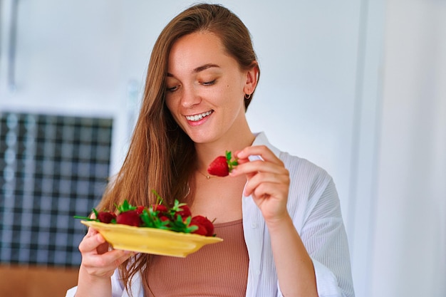 Linda garota feliz sorridente satisfeita comendo morangos maduros frescos na cozinha de casa