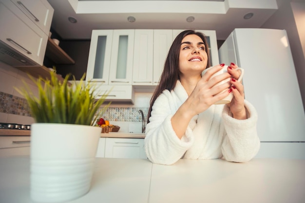 Linda garota feliz usa roupão branco toma café na cozinha pela manhã