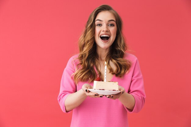 Linda garota feliz, vestindo roupas casuais, em pé, isolada na parede rosa, comemorando aniversário com bolo