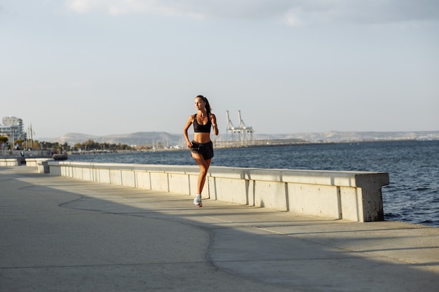 Linda garota fitness com shorts e top esportivo corre à beira-mar à beira-mar durante o pôr do sol